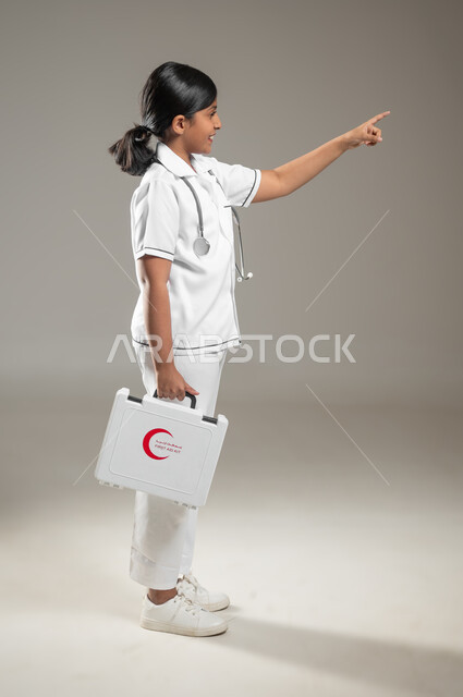 مبادئ التعامل مع حالات الطوارئ، النظر إلى الكاميرا، بورتريه من الجانب لفتاة عربية خليجية سعودية ترتدي اللباس الطبي وتحمل بيدها حقيبة الاسعافات الأولية تشير الى مكان ما، الجسم بالطول الكامل، خلفية رمادية، محتوى حساس، استخدام حساس