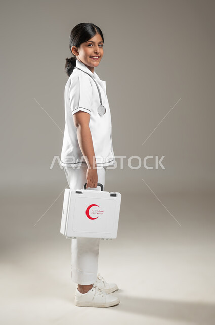 النظر إلى الكاميرا، آمال مستقبلية في الوصول للحلم، بورتريه من الجانب لفتاة عربية خليجية سعودية ترتدي اللباس الطبي وتحمل بيدها حقيبة الاسعافات الأولية، مساعدة المرضى وطريقة التعامل في الحالات الطارئة، الجسم بالطول الكامل، خلفية رمادية، محتوى حساس، استخدام حساس