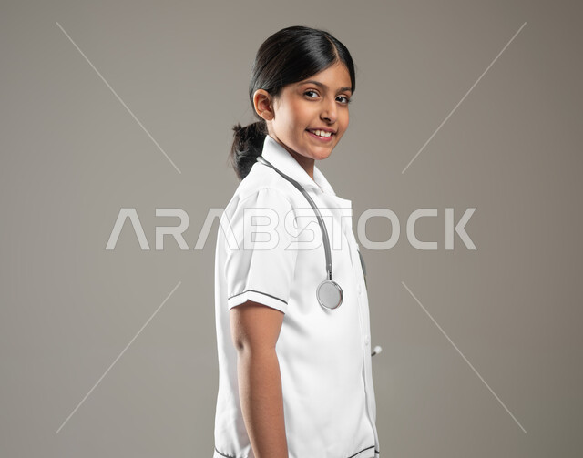 النظر الى الكاميرا بايماءات البشاشه، الاهتمام بتقديم الخدمات الطبية والاستعداد لحالات الطوارئ,  بورتريه مقرب من الجانب لفتاة عربية خليجية سعودية ترتدي اللباس الطبي مع السماعة الطبية ، خلفية رمادية، محتوى حساس، استخدام حساس