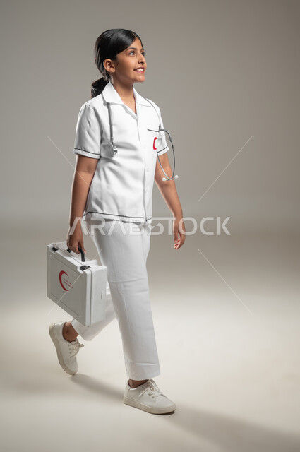 آحلام مستقبلية في الوصول للحلم، النظر إلى الكاميرا، بورتريه من الجانب لفتاة عربية خليجية سعودية ترتدي اللباس الطبي وتحمل بيدها حقيبة الاسعافات الأولية، مساعدة المرضى وطريقة التعامل في الحالات الطارئة، الجسم بالطول الكامل، خلفية رمادية، محتوى حساس، استخدام حساس