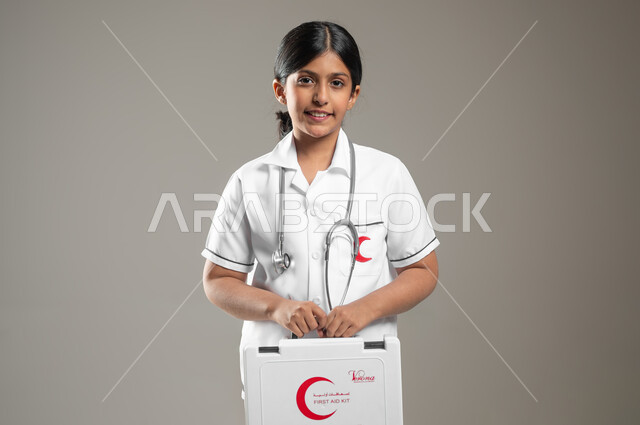 انقاذ المرضى في الحالات الطارئة، الأهداف المستقبلية في العمل في المستشفيات، بورتريه مقرب لفتاة عربية خليجية سعودية مبتسمة ترتدي اللباس الطبي تحمل بيدها حقيبة الاسعافات الاولية تحلم أن تصبح طبيبة، خلفية رمادية، محتوى حساس، استخدام حساس