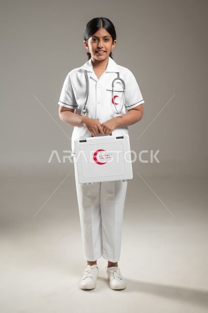مساعدة المرضى في الحالات الطارئة، الأهداف المستقبلية في العمل في المستشفيات، بورتريه لفتاة عربية خليجية سعودية مبتسمة ترتدي اللباس الطبي تحمل بيدها حقيبة الاسعافات الاولية تحلم أن تصبح طبيبة, الجسم بالطول الكامل، خلفية رمادية، محتوى حساس، استخدام حساس