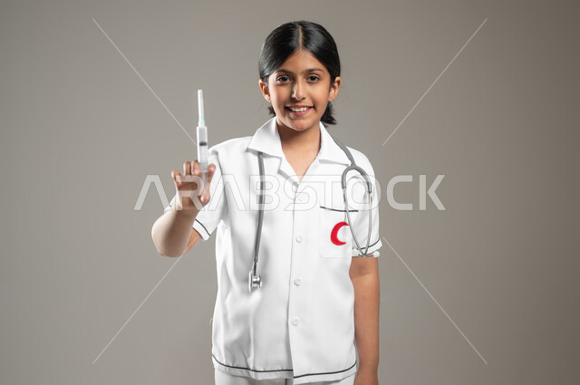 آمال مستقبيله في العمل في المختبرات والتحاليل، ايماءات الشغف بالحصول على الشهادات العليا، مفهوم الطب والرعاية الصحية، بورتريه لفتاة عربية خليجية سعودية مبتسمة ترتدي اللباس الطبي وتحمل بيدها حقنة طبية، الجسم بالطول الكامل، خلفية رمادية، محتوى حساس، استخدام حساس