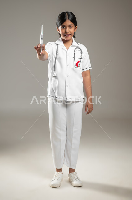 ايماءات الشغف بالحصول على الشهادات العليا، آمال مستقبيله في العمل في المختبرات والتحاليل ،مفهوم الطب والرعاية الصحية، بورتريه لفتاة عربية خليجية سعودية مبتسمة ترتدي اللباس الطبي وتحمل بيدها حقنة طبية، الجسم بالطول الكامل، خلفية رمادية، محتوى حساس، استخدام حساس