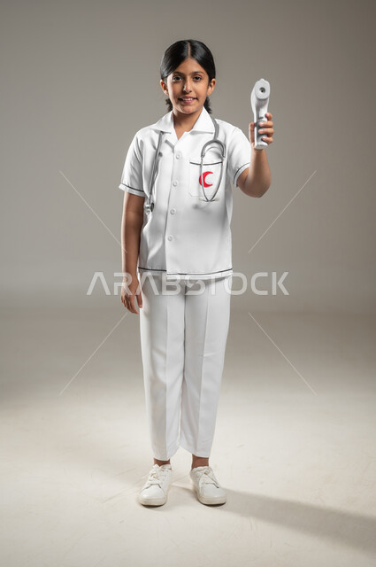 أهداف مستقبلية في العمل في المستشفيات، تحديد مهنة المستقبل منذ الصغر، اتباع إجراءات السلامة والوقاية، بورتريه لفتاة عربية خليجية سعودية مبتسمة ترتدي اللباس الطبي تحمل بيدها ميزان الحرارة الرقمي، الجسم بالطول الكامل، خلفية رمادية، محتوى حساس، استخدام حساس