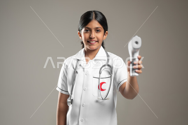 Measuring temperature using a thermometer, determining a future profession from a young age, following safety and prevention measures, portrait of a smiling Saudi Arabian Gulf girl wearing a medical uniform holding a digital thermometer in her hand, gray background, sensitive content, sensitive use