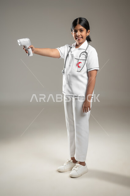 تحديد مهنة المستقبل منذ الصغر، اتباع إجراءات السلامة والوقاية، أهداف مستقبلية في العمل في المستشفيات، بورتريه لفتاة عربية خليجية سعودية مبتسمة ترتدي اللباس الطبي تحمل بيدها ميزان الحرارة الرقمي، الجسم بالطول الكامل، خلفية رمادية، محتوى حساس، استخدام حساس