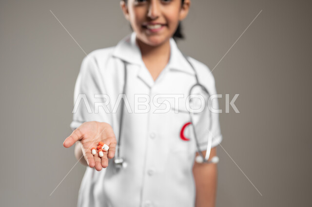 الالتزام بالأوقات الصحيحة لأخذ العلاج, استخدام عقار الدواء بوصفة طبية, بورتريه مقرب لفتاة عربية خليجية سعودية مبتسمة ترتدي اللباس الطبي تمسك حبوب الدواء, مفهوم الطب والرعاية الطبية, خلفية رمادية، محتوى حساس، استخدام حساس
