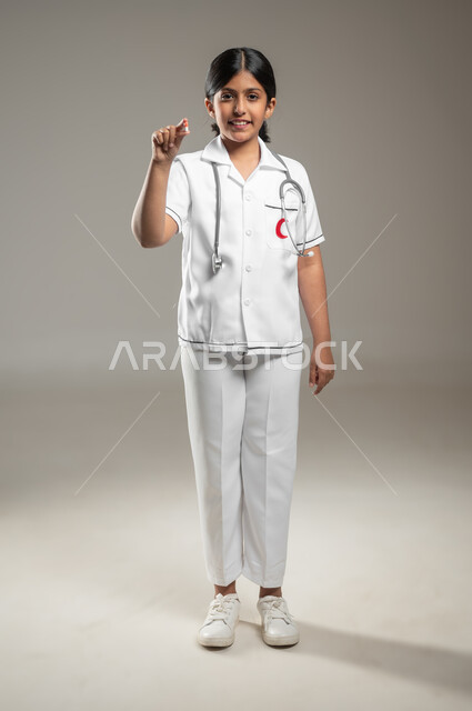 الاشارة إلى الكاميرا بإيماءات الفرح والسرور، الطموحات و الأهداف المستقبلية في العمل في المستشفيات، بورتريه لفتاة عربية خليجية سعودية مبتسمة ترتدي اللباس الطبي تحلم أن تصبح طبيبة إسعافات أولية، خلفية رمادية، الجسم بالطول الكامل، محتوى حساس، استخدام حساس