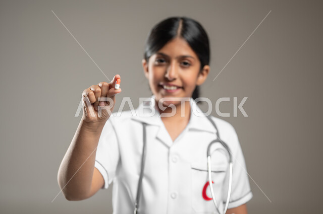 Determining the profession since childhood, pointing to the camera with gestures of joy and pleasure, ambitions and future goals in working in hospitals, a close-up portrait of a smiling Saudi Arabian Gulf girl wearing a medical uniform dreaming of becoming a first aid doctor, gray background, sensitive content, sensitive use