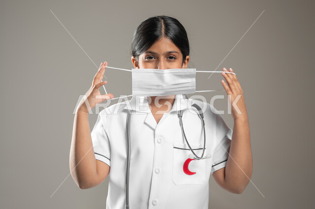 Avoiding the spread of Covid 19, wearing a mask to prevent the Corona virus, health awareness and avoiding the spread of infection, a close-up portrait of a Saudi Arabian Gulf girl wearing a medical gown and a mask, adhering to preventive precautions, gray background, sensitive content, sensitive use