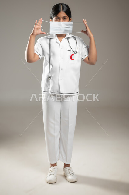 الحفاظ على السلامة و تجنب انتشار العدوى من كوفيد 19, ارتداء الكمامه للوقاية من فايروس كورونا، بورتريه مقرب لفتاة عربية خليجية سعودية ترتدي اللباس الطبي و الكمامة، الجسم بالطول الكامل، خلفية رمادية، محتوى حساس، استخدام حساس