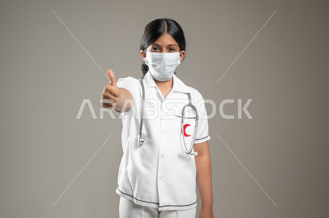 Health awareness and avoiding the spread of infection, the concept of medicine and health care, a close-up portrait of a Saudi Gulf Arab girl wearing a medical gown and a mask, pointing with her thumb up with different facial and hand gestures indicating admiration, gray background, sensitive content, sensitive use