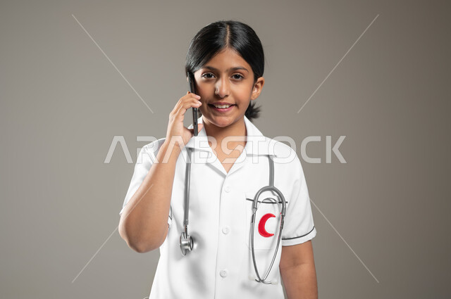 Future ambitions and goals for working in hospitals, using technology for medical consultations, a close-up portrait of a Saudi Gulf Arab girl wearing medical clothing making a phone call via mobile phone, gray background, sensitive content, sensitive use