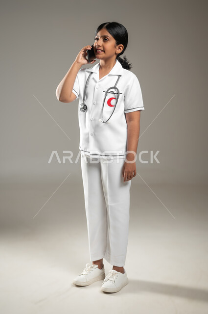 استخدام التكنولوجيا للإستشارات الطبية ،تلبية مطالب الرعاية الصحية باستخدام التقنيات الحديثة، بورتريه لفتاه عربية خليجية سعودية ترتدي اللباس الطبي تقوم بإجراء مكالمة هاتفيه عبر الهاتف المحمول، الجسم بالطول الكامل، خلفية رمادية، محتوى حساس، استخدام حساس