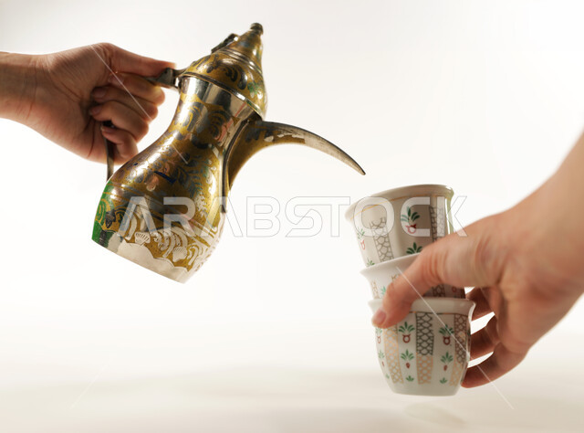 حسن الضيافه والإستقبال في الأعياد والمناسبات الدينية، منتجات وطنية محلية، الاستمتاع بالمشروبات الساخنة، اتباع العادات والتقاليد السعودية في تقديم القهوة العربية بالدله النحاسية والفنجان، صورة مقربة ليد رجل عربي حليجي يقوم بصب القهوة