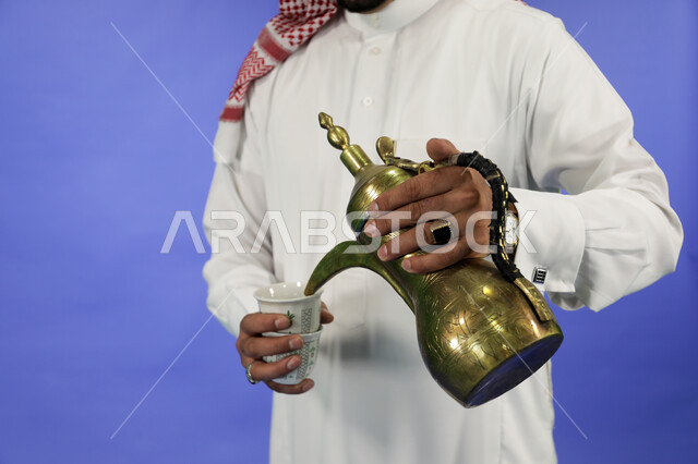 اتباع العادات والتقاليد السعودية في تقديم القهوة العربية بالدله النحاسية والفنجان، حسن الضيافه والإستقبال في الأعياد والمناسبات الدينية، منتجات وطنية محلية، الاستمتاع بالمشروبات الساخنة، صورة مقربة ليد رجل عربي حليجي يرتدي الثوب التقليدي والشماغ يقوم بصب القهوة