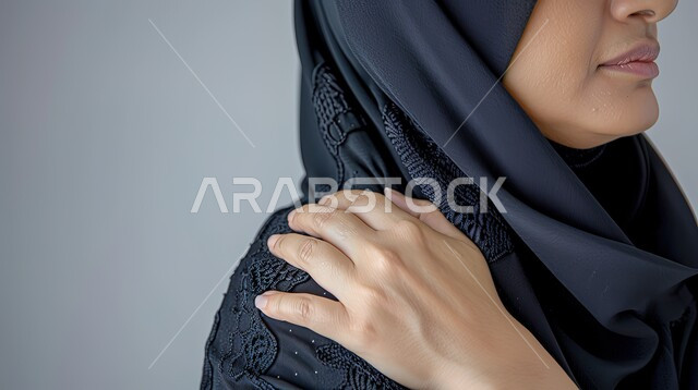 Inflammation of muscles and joints and the need for treatment, suffering from nervous and muscular aches and pains, close-up portrait of a Saudi Arabian Gulf woman wearing an abaya and hijab, placing her hand on her shoulder with gestures of pain, fatigue and exhaustion, gray background.