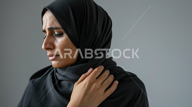 الأوجاع والآلام العصبية والعضلية، طلب المساعده والحاجه للعلاج، التهاب العضلات والمفاصل، بورتريه مقرب لإمرأة عربيه سعوديه خليجيه ترتدي العباءة والحجاب تضع يدها على كتفها بإيماءات الألم والتعب والأرهاق, خلفية رمادية