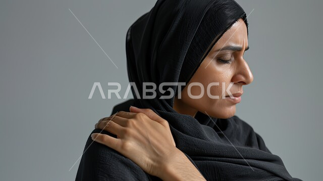 طلب المساعده والحاجه للعلاج، الأوجاع والآلام العصبية والعضلية، التهاب العضلات والمفاصل، بورتريه مقرب لإمرأة عربيه سعوديه خليجيه ترتدي العباءة والحجاب تضع يدها على كتفها بإيماءات الألم والتعب والأرهاق, خلفية رمادية