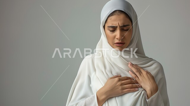 الذبحه الصدريه حالة مرضية، مشاكل صحية صعبة، إيماءات التعب والإرهاق والضغوطات النفسية، بورتريه مقرب لإمرأة عربية سعودية خليجية محجبة ترتدي العباءة  تضع يدها على قلبها تشعر بحالة سيئة من الوخز والألم, خلفية رمادية