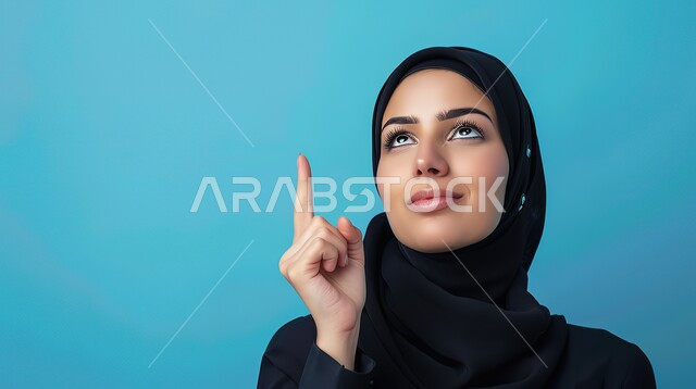 حركات وإشارات يدين تدل على هذا المكان (هنا)، تحديد موقع معين بإيماءات البشاشة، بورتريه مقرب لامرأة عربيه خليجيه سعوديه مبتسمه محجبه ترتدي عباءة سوداء تقوم بالنظر الى الأعلى وترفع اصابعها السبابة وتشير الى شيء ما، خلفية زرقاء