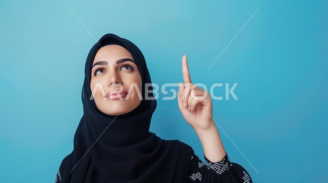 تحديد موقع معين بإيماءات البشاشة، إشارات وحركات يدين تدل على هذا المكان (هنا)، بورتريه مقرب لامرأة عربيه خليجيه سعوديه مبتسمه محجبه ترتدي عباءة سوداء تقوم بالنظر الى الأعلى وترفع اصابعها السبابة وتشير الى شيء ما، خلفية زرقاء