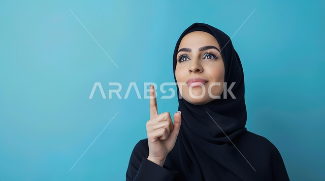 إشارات وحركات يدين تدل على هذا المكان (هنا)، تحديد موقع معين بإيماءات البشاشة، بورتريه مقرب لامرأة عربيه خليجيه سعوديه مبتسمه محجبه ترتدي عباءة سوداء تقوم بالنظر الى الأعلى وترفع اصابعها السبابة وتشير الى شيء ما، خلفية زرقاء
