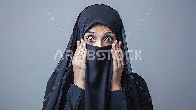 التعجب والصدمة من حصول أمر ما، النظر إلى الكاميرا بتعابير الدهشة والخوف، بورتربه مقرب لامرأة عربية خليجية سعودية محجبة ترتدي عباءة سوداء تضع يديها على وجهها بإيماءات تدل على الذهول، خلفية رمادية