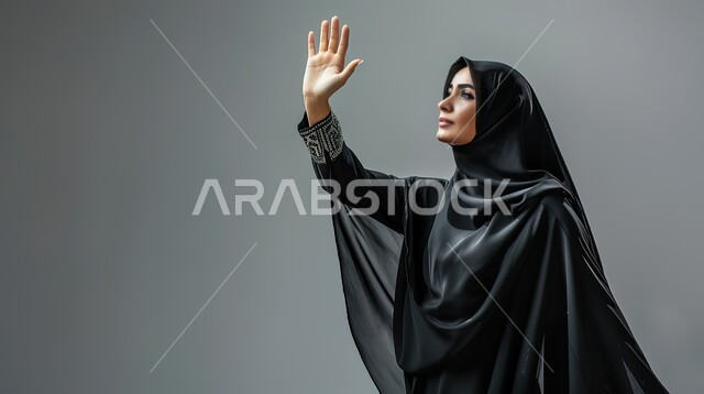 تعابير الترحيب والاستقبال، ايماءات إلقاء التحية والسلام، بورتريه مقرب لإمرأة عربية خليجية سعودية محجبة ترتدي عباءة سوداء تقوم برفع يدها للأعلى، خلفية رمادية
