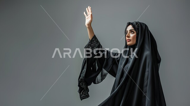 ايماءات إلقاء التحية والسلام، تعابير الترحيب والاستقبال، بورتريه مقرب لإمرأة عربية خليجية سعودية محجبة ترتدي عباءة سوداء تقوم برفع يدها للأعلى، خلفية رمادية