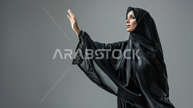ايماءات إلقاء التحية والسلام، تعابير الترحيب والاستقبال، بورتريه مقرب لإمرأة عربية خليجية سعودية محجبة ترتدي عباءة سوداء تقوم برفع يدها للأعلى، خلفية رمادية
