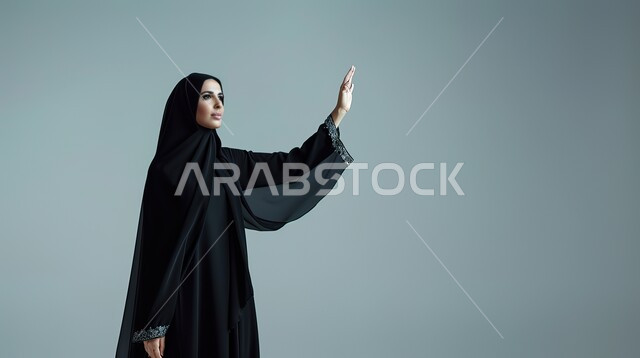 تعابير الترحيب والاستقبال، ايماءات إلقاء التحية والسلام، بورتريه مقرب لإمرأة عربية خليجية سعودية محجبة ترتدي عباءة سوداء تقوم برفع يدها للأعلى، خلفية رمادية