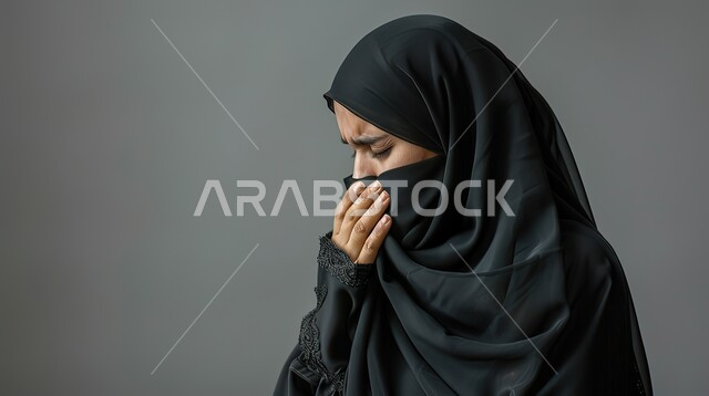 الشعور بالتعب والمرض، الإصابة بفايروس كورونا كوفيد19، بورتريه مقرب من الجانب لامرأه عربيه خليجيه سعوديه منقبة ترتدي عباءة سوداء، إيماءات وجه ويدين تدل على العطاس، خلفية رمادية