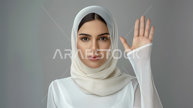 الشعور بالإنزعاج والغضب، الأمر بالتوقف عن فعل شيء ما، تعابير الرفض والإمتناع، بورتريه مقرب لامرأه عربيه خليجيه سعوديه محجة ترتدي عباءة بيضاء ترفع يدها بإيماءات تدل على النهي، خلفية رمادية