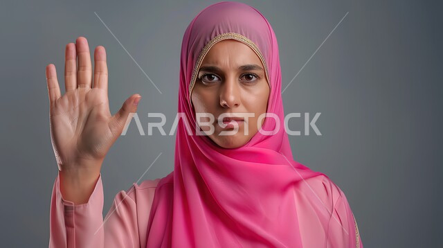 تعابير الرفض والإمتناع، الأمر بالتوقف عن فعل شيء ما، بورتريه مقرب لامرأه عربيه خليجيه سعوديه محجة ترتدي عباءة وردية ترفع يدها بإيماءات تدل على النهي، الشعور بالإنزعاج والغضب، خلفية رمادية