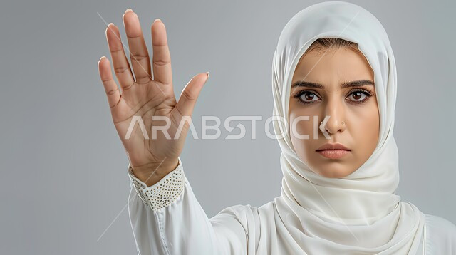 الشعور بالإنزعاج والغضب، الأمر بالتوقف عن فعل شيء ما، تعابير الرفض والإمتناع، بورتريه مقرب لامرأه عربيه خليجيه سعوديه محجة ترتدي عباءة بيضاء ترفع يدها بإيماءات تدل على النهي، خلفية رمادية