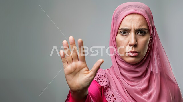 الأمر بالتوقف عن فعل شيء ما، تعابير الرفض والإمتناع، بورتريه مقرب لامرأه عربيه خليجيه سعوديه محجة ترتدي عباءة وردية ترفع يدها بإيماءات تدل على النهي، الشعور بالإنزعاج والغضب، خلفية رمادية