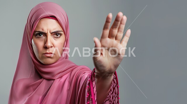 تعابير الرفض والإمتناع، الأمر بالتوقف عن فعل شيء ما، بورتريه مقرب لامرأه عربيه خليجيه سعوديه محجة ترتدي عباءة وردية ترفع يدها بإيماءات تدل على النهي، الشعور بالإنزعاج والغضب، خلفية رمادية