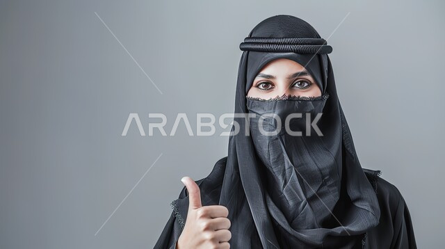 إيماءات الامتياز والجودة، مفهوم القبول والموافقه، بورتريه مقرب لامرأه عربيه خليجيه سعوديه منقبة ترتدي عباءة سوداء ترفع اصبع الابهام بتعابير الإعجاب والرضى، خلفية رمادية