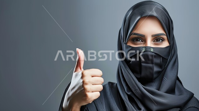 مفهوم القبول والموافقه، تعابير الإعجاب والرضى، بورتريه مقرب لامرأه عربيه خليجيه سعوديه منقبة ترتدي عباءة سوداء ترفع اصبع الابهام بإيماءات الامتياز والجودة، خلفية رمادية