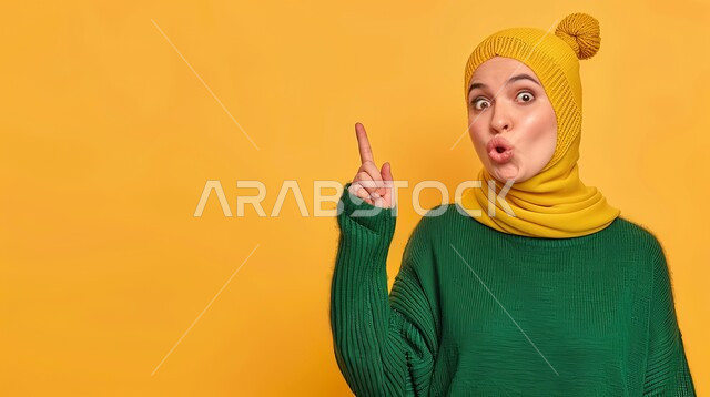 تعابير السرور والفرح، تحديد موقع معين بإيماءات البشاشة، إشارات وحركات يدين تدل على هذا المكان (هنا)، بورتريه مقرب لامرأة عربيه خليجيه سعوديه مبتسمه محجبه ترتدي الزي الكاجوال ترفع اصابعها السبابة وتشير الى شيء ما، خلفية صفراء