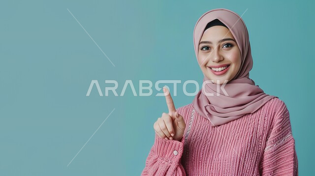 إشارات وحركات يدين تدل على هذا المكان (هنا)، تحديد موقع معين بإيماءات البشاشة، بورتريه مقرب لامرأة عربيه خليجيه سعوديه مبتسمه محجبه ترتدي الزي الكاجوال ترفع اصابعها السبابة وتشير الى شيء ما، خلفية زرقاء