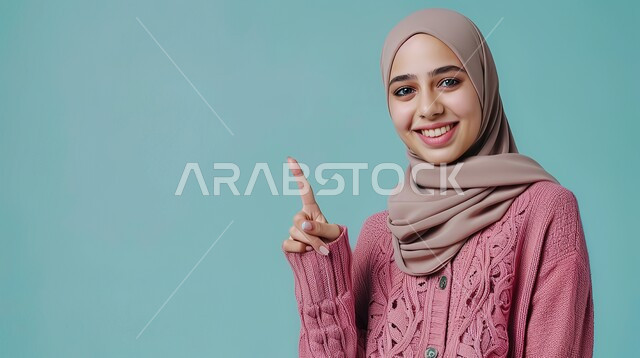 تعابير السرور والفرح، تحديد موقع معين بإيماءات البشاشة، إشارات وحركات يدين تدل على هذا المكان (هنا)، بورتريه مقرب لامرأة عربيه خليجيه سعوديه مبتسمه محجبه ترتدي الزي الكاجوال ترفع اصابعها السبابة وتشير الى شيء ما، خلفية زرقاء