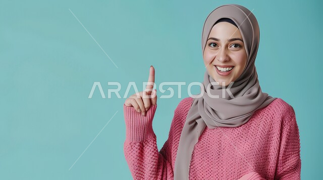 إشارات وحركات يدين تدل على هذا المكان (هنا)، تحديد موقع معين بإيماءات البشاشة، بورتريه مقرب لامرأة عربيه خليجيه سعوديه مبتسمه محجبه ترتدي الزي الكاجوال ترفع اصابعها السبابة وتشير الى شيء ما، خلفية زرقاء