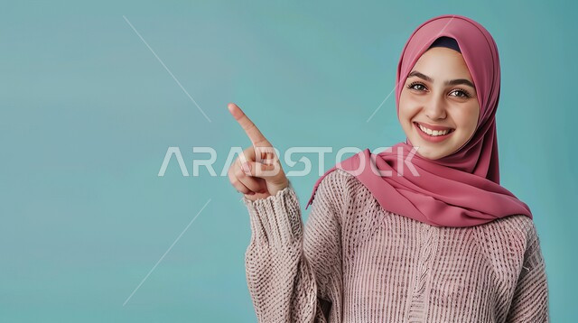 تحديد موقع معين بإيماءات البشاشة، إشارات وحركات يدين تدل على هذا المكان (هنا)، بورتريه مقرب لامرأة عربيه خليجيه سعوديه مبتسمه محجبه ترتدي الزي الكاجوال ترفع اصابعها السبابة وتشير الى شيء ما، خلفية زرقاء