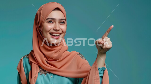 تعابير السرور والفرح، تحديد موقع معين بإيماءات البشاشة، إشارات وحركات يدين تدل على هذا المكان (هنا)، بورتريه مقرب لامرأة عربيه خليجيه سعوديه مبتسمه محجبه ترتدي الزي الكاجوال ترفع اصابعها السبابة وتشير الى شيء ما، خلفية زرقاء