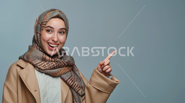 تحديد موقع معين بإيماءات البشاشة، إشارات وحركات يدين تدل على هذا المكان (هنا)، بورتريه مقرب لامرأة عربيه خليجيه سعوديه مبتسمه محجبه ترتدي الزي الكاجوال ترفع اصابعها السبابة وتشير الى شيء ما، خلفية رمادية