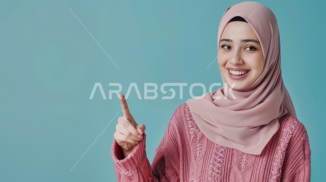 إشارات وحركات يدين تدل على هذا المكان (هنا)، تحديد موقع معين بإيماءات البشاشة، بورتريه مقرب لامرأة عربيه خليجيه سعوديه مبتسمه محجبه ترتدي الزي الكاجوال ترفع اصابعها السبابة وتشير الى شيء ما، خلفية زرقاء