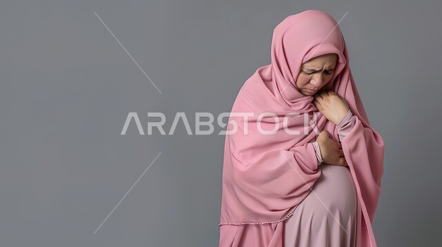 الحاجة لوضع خطة علاجية، حرقة في المعدة وعسر الهضم، الشعور بالشعور بآلام المخاض  وقرب الولاده، الإرهاق والتعب والإحساس بالمغص، بورتريه مقرب لإمرأة عربية سعودية خليجية محجبة حامل ترتدي العباءة تمسك بطنها بإيماءات الشعور بالألم، خلفية رمادية